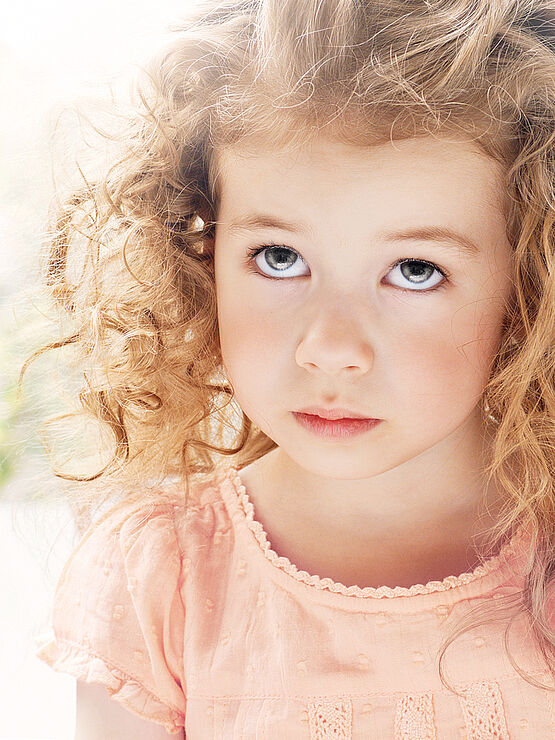 Young girl with curly hair looking up with a serious expression, wearing a pink dress, in a softly lit environment